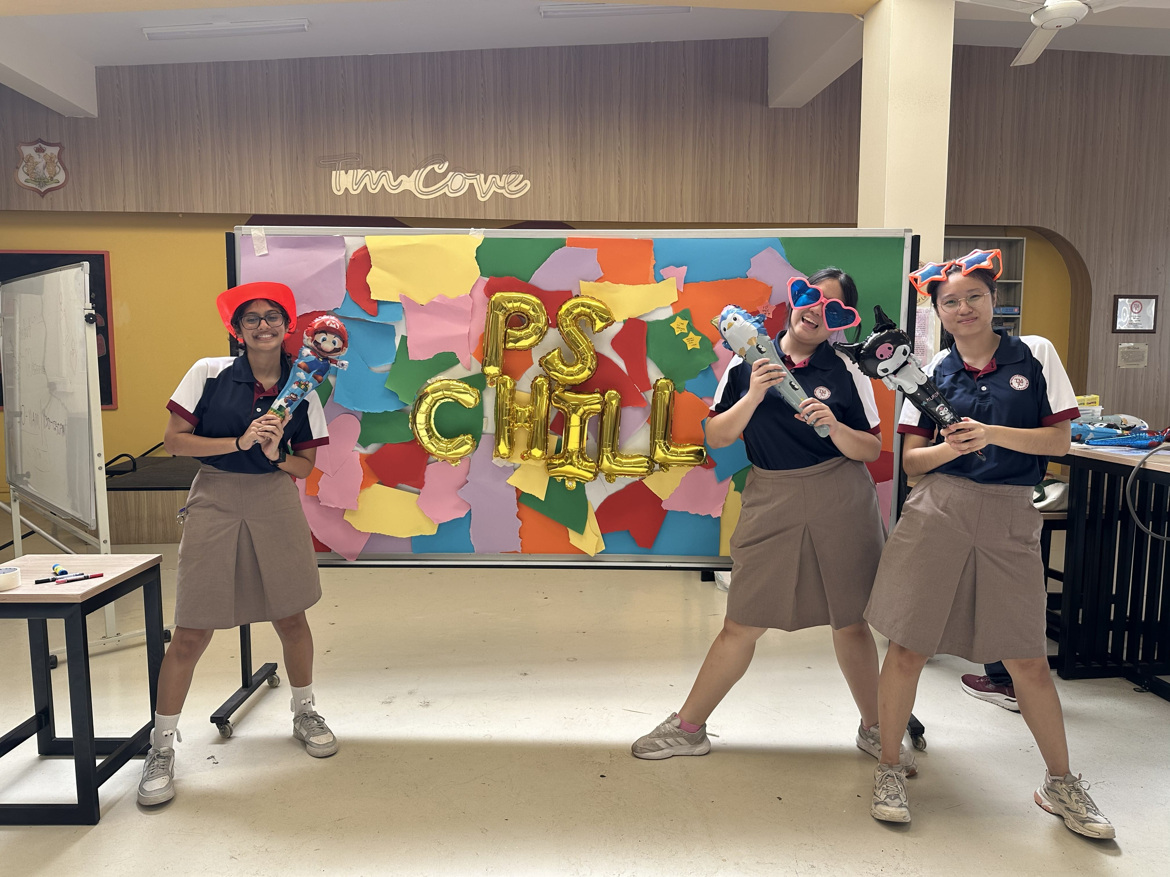 Three female students in school uniforms pose playfully in front of a colourful paper collage backdrop with gold balloon letters spelling "PS CHILL", holding toy props and wearing fun accessories.